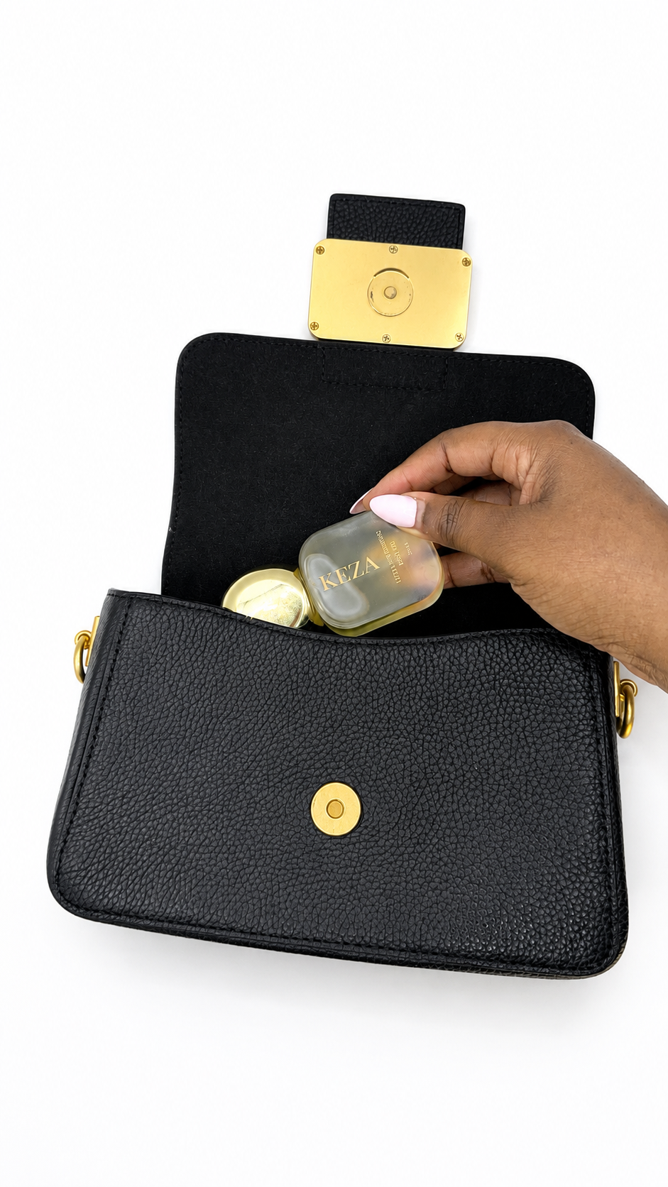 A black textured leather crossbody bag with gold-tone hardware, shown open with a hand placing a small bottle of KEZA body oil inside. The bag has a black flap with a gold clasp and a gold snap button on the front.